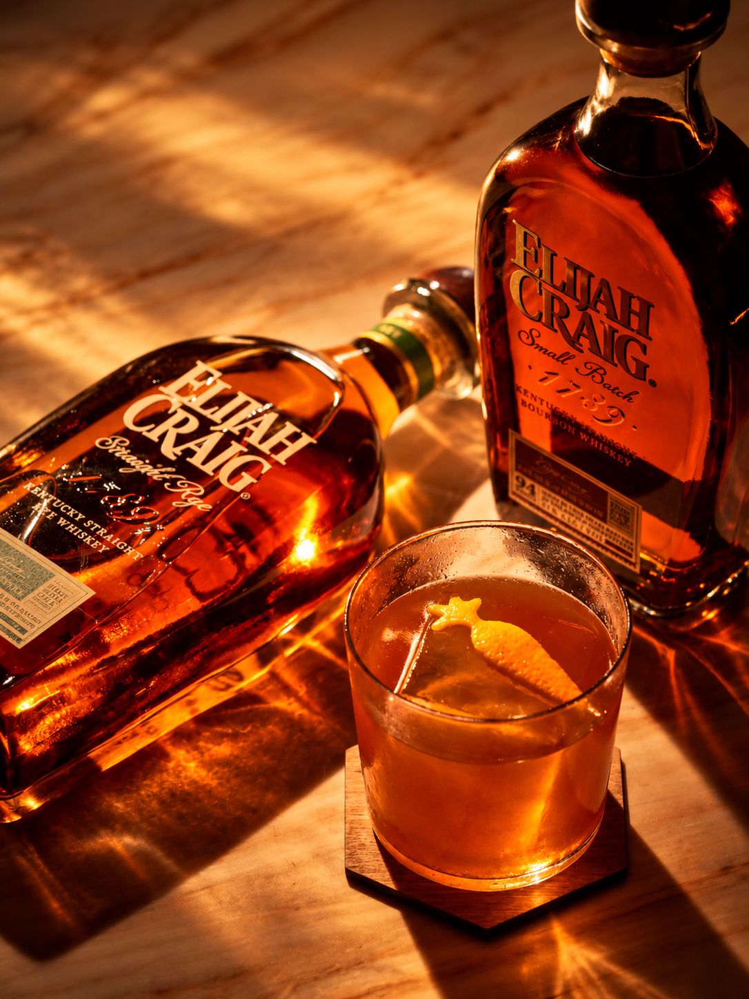 Old Fashioned Week Bruxelles 2026 : Elijah Craig s’invite dans cinq bars bruxellois Deux bouteilles d’Elijah Craig et un Old Fashioned garni d’un zeste d’orange sur un comptoir en bois éclairé d’une lumière ambrée