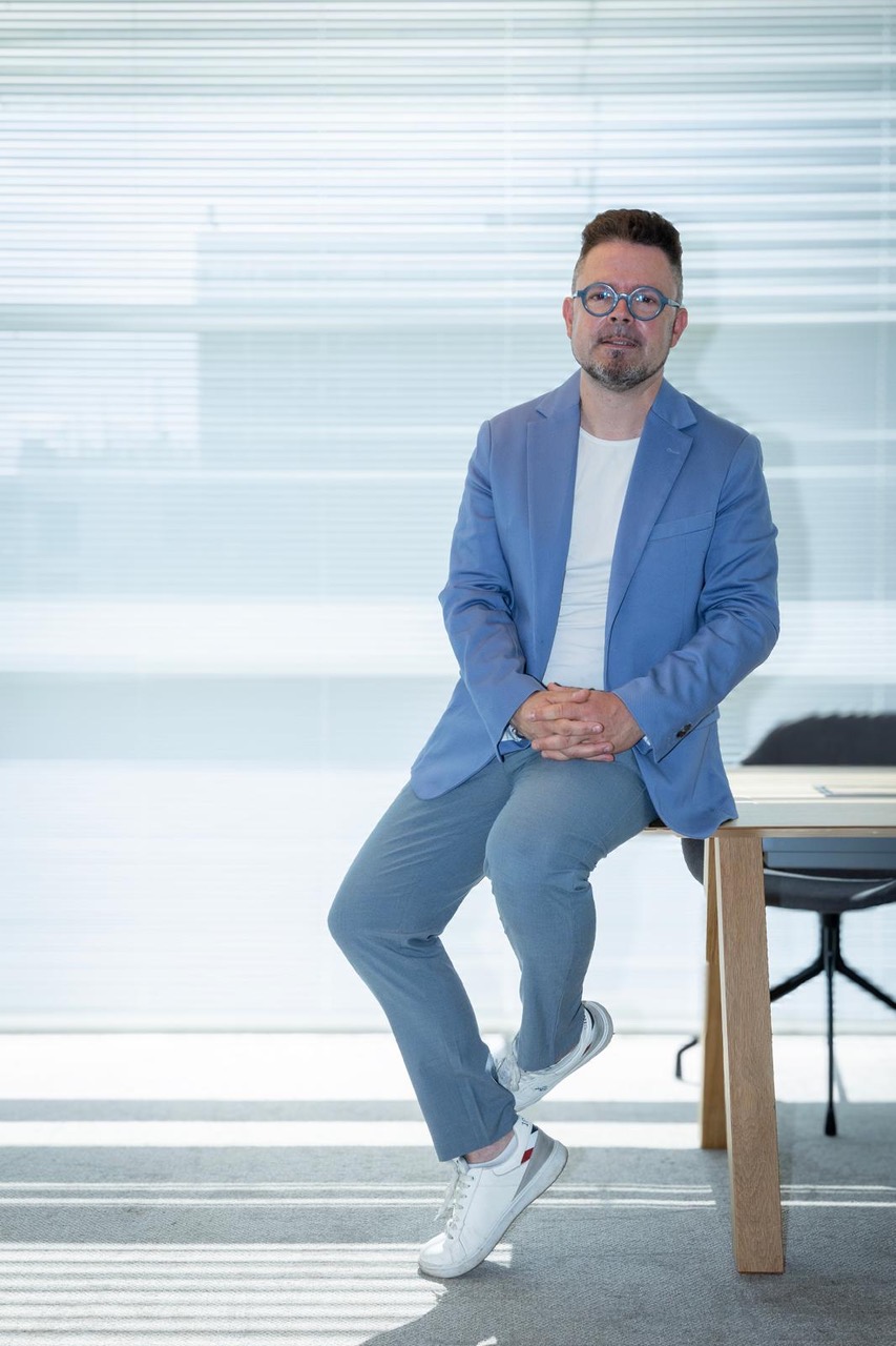 Portret van een man in een blauw jasje, zittend op een tafel in een licht kantoor