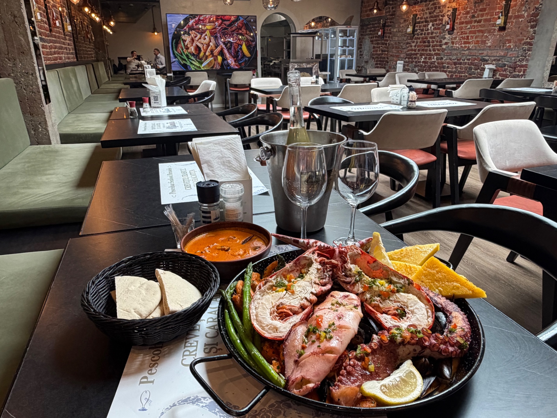 Table dressée avec plateau de homard et poulpe, bisque de homard, polenta dorée, bouteille de vin blanc en seau à glace, vue sur la salle