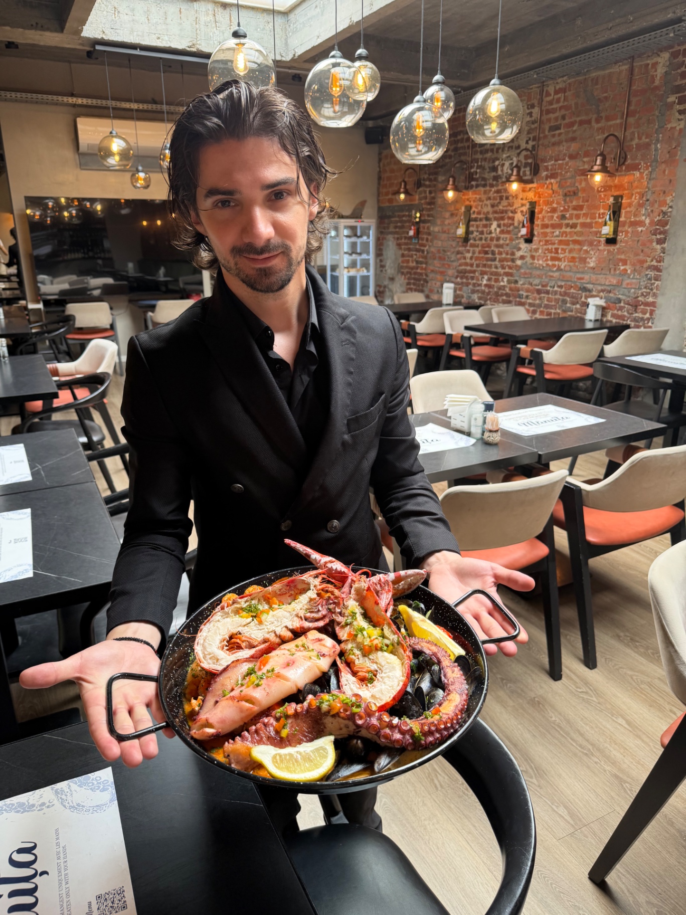 © Bernard Frisque pour BXLove · Pescobar Seafood Brussels, juin 2026 Maître d'hôtel de Pescobar Brussels présentant un plateau seafood signature — homard, poulpe, gambas, moules — sous les suspensions en verre de la salle aux murs de briques