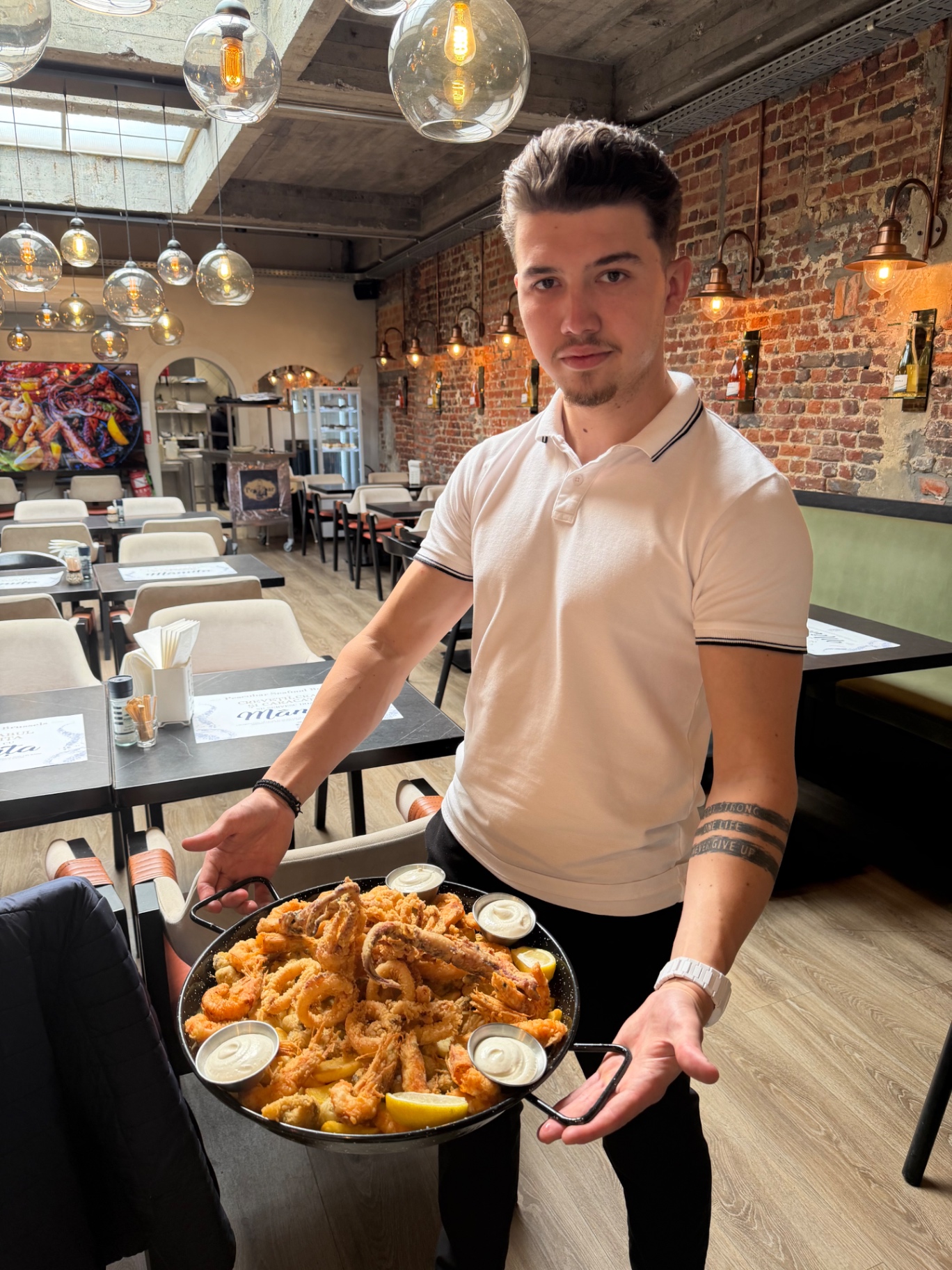 Serveur Pescobar présentant un Fritto Misto géant dans une paella : crevettes, calamars, poissons frits dorés, quartiers de citron, sauces tartare