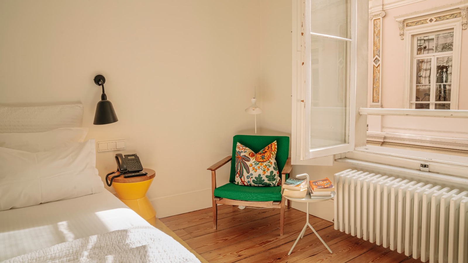 Chambre lumineuse de l’Hôtel des Galeries, au cœur des Galeries Royales Saint-Hubert. Lichte kamer van Hôtel des Galeries met houten vloer, groene fauteuil en open raam op een historische Brusselse gevel