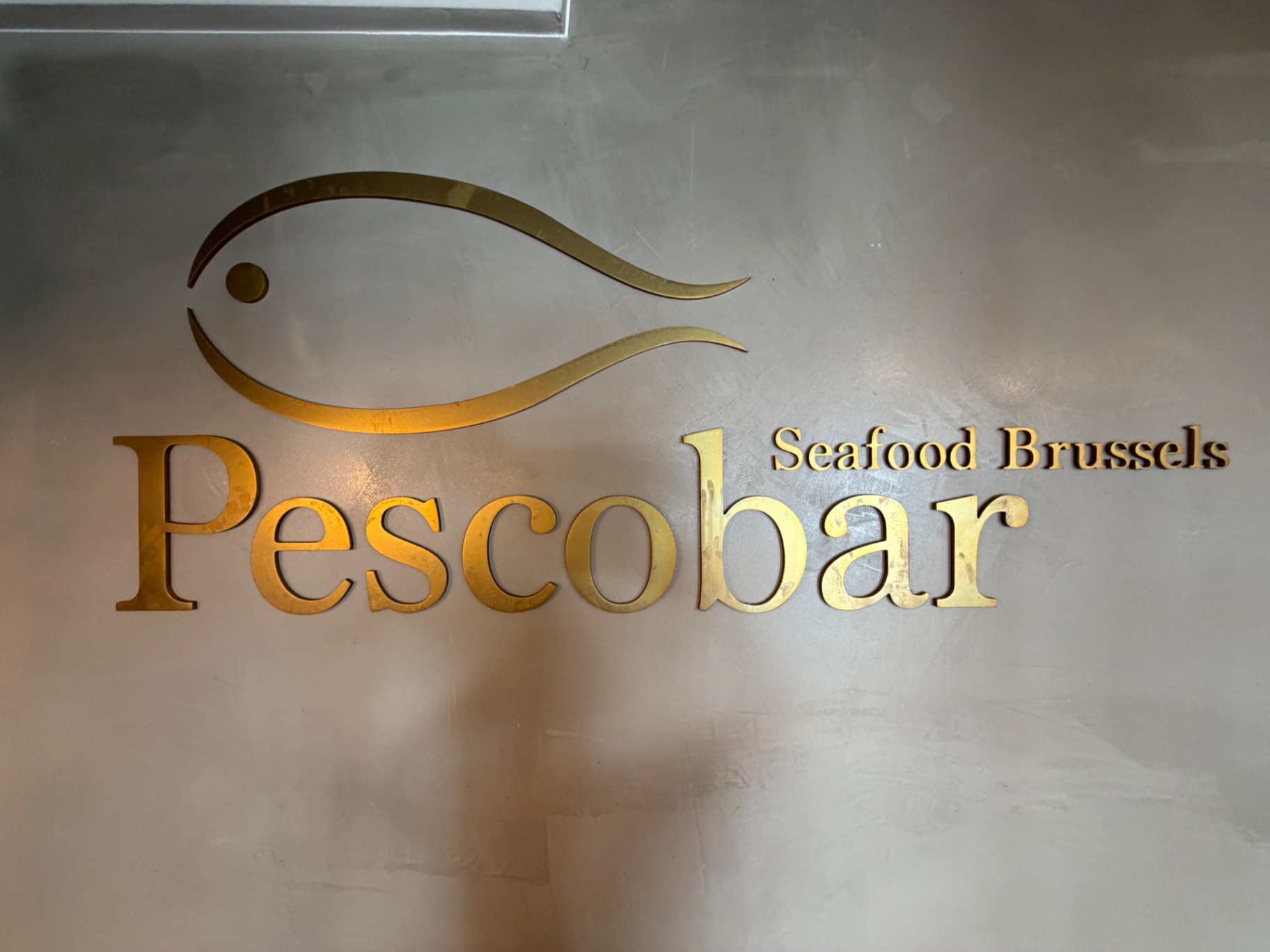 Logo doré Pescobar Seafood Brussels sur mur béton ciré — poisson stylisé en lettres dorées